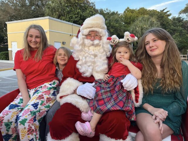 Santa sitting on the sleigh with four smiling kids, one in a plaid dress on his lap