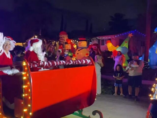Santa and Mrs. Claus waving from the lit-up red sleigh past inflatable decorations at night
