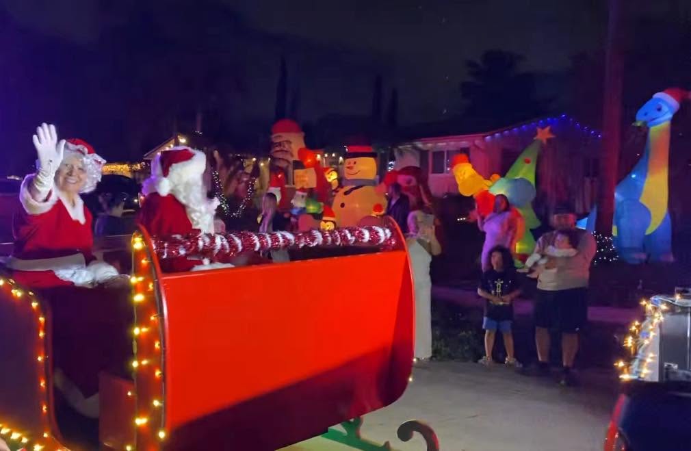 Santa and Mrs. Claus waving from the lit-up red sleigh past inflatable decorations at night