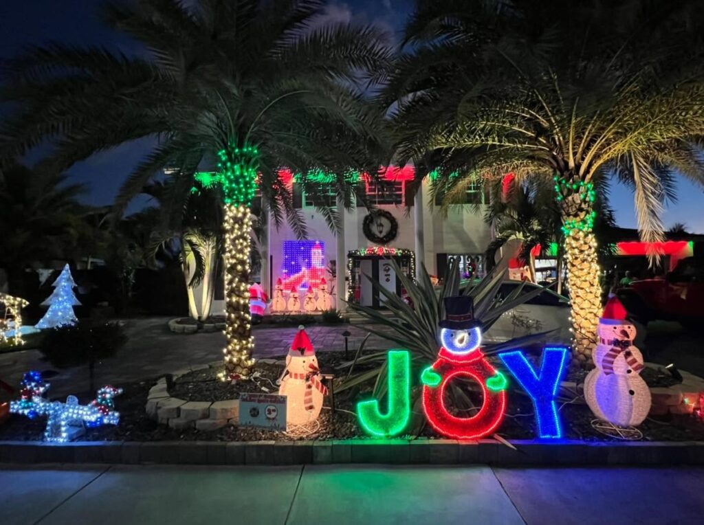 A front yard with a glowing JOY sign, a snowman, palm trees wrapped in lights and an inflatable Santa
